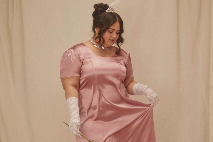 kobieta ubrana w suknię regencyjną oraz białe rękawiczki balowe i pokazująca na beżowym tle, tworząc sesję zdjęciową jak z Bridgertonów