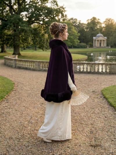 kobieta idącą ścieżką w parku ubrana w białą suknię regencyjną, ciemnofioletową elegancką pelerynę damską oraz trzymająca biały wachlarz