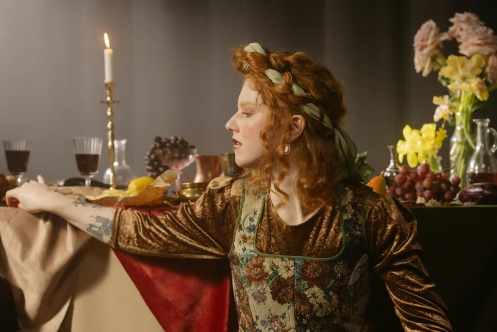 kobieta z rudymi włosami ubrana w brązową sukienkę oraz gorset w kwiaty, opierająca się o stół zastawiony jedzeniem