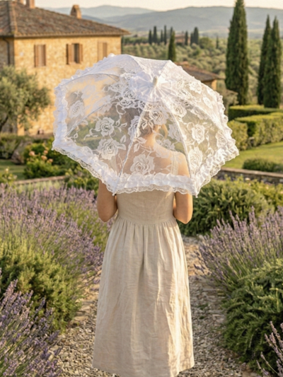 kobieta w beżowej sukience idąca tyłem kamienną ścieżką trzymająca białą parasolkę koronkową