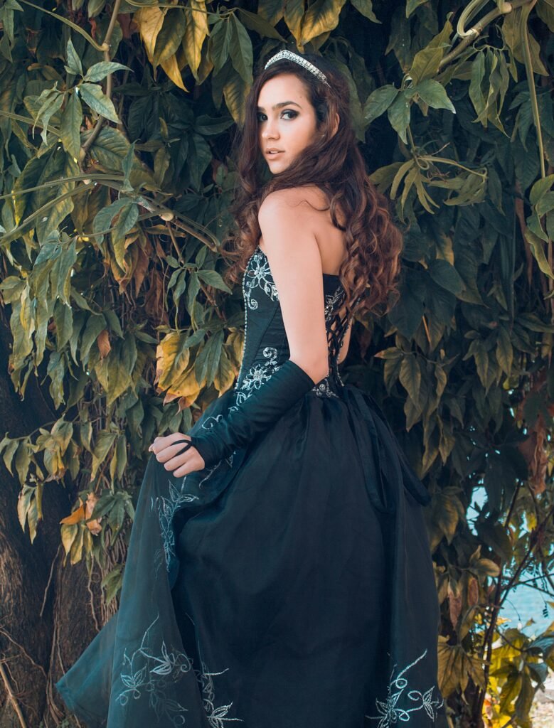 kobieta z ciemnymi falowanymi włosami nosząca czarną suknię balową oraz srebrną tiarę, liście drzewa w tle